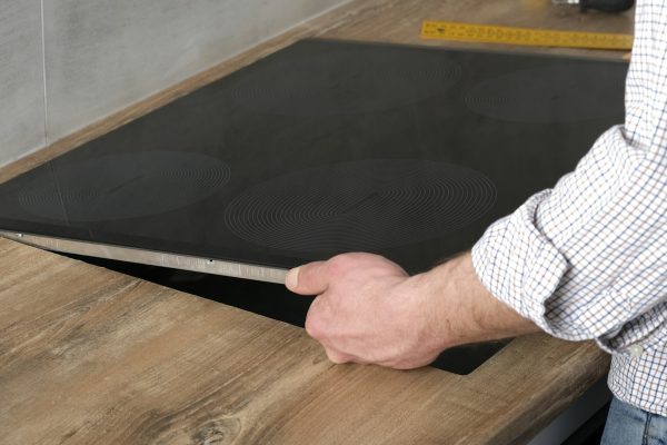 A young repairman installs a black induction hob in a modern white Scandinavian style kitchen A young repairman installs a black induction hob in a modern white Scandinavian style kitchen